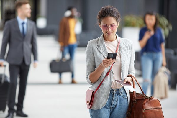 Smiling beautiful young woman in sunglasses carrying travel bag and using mobile internet in airport