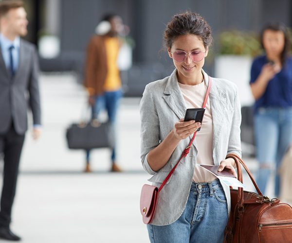 Smiling beautiful young woman in sunglasses carrying travel bag and using mobile internet in airport